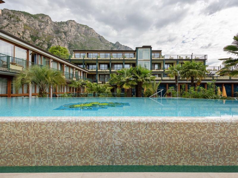 Moderner Hotelpool mit klar blauem Wasser, umgeben von Palmen und Gebäuden unter bewölktem Himmel.