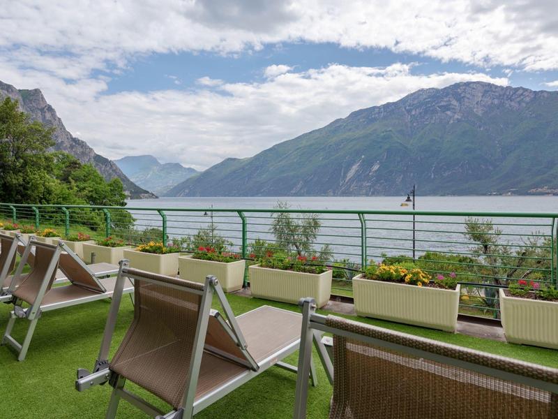 Grüner Balkon mit Liegestühlen und Blumen vor Bergsee und bewölktem Himmel.