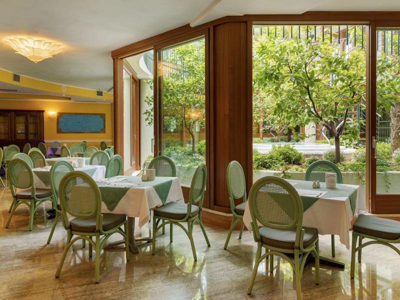 Helles Restaurant mit großen Fenstern und grünen Stühlen, Blick auf Garten mit Bäumen draußen.