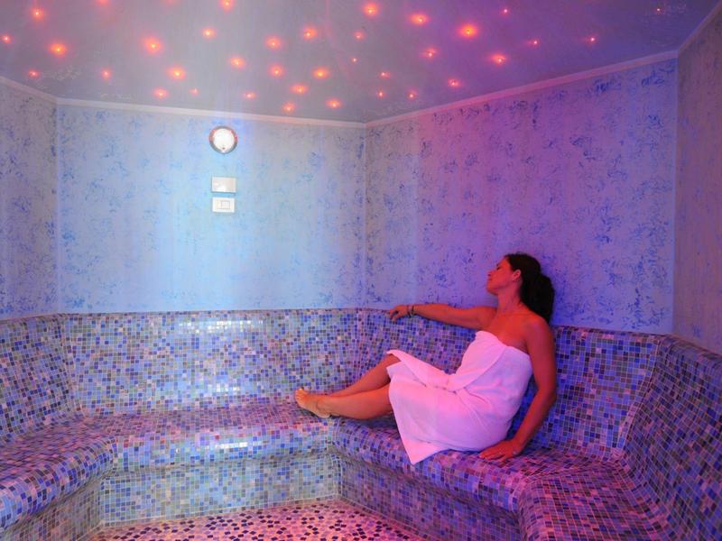 Frau mit Handtuch sitzt entspannt in dampfigem Dampfbad mit bunter Deckenbeleuchtung.