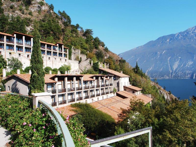 Budynek hotelu na zalesionym zboczu z widokiem na jezioro i góry w tle.