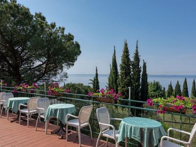 Terrasse mit Tischen und Stühlen, blauer Himmel, Zypressen und Meer im Hintergrund.