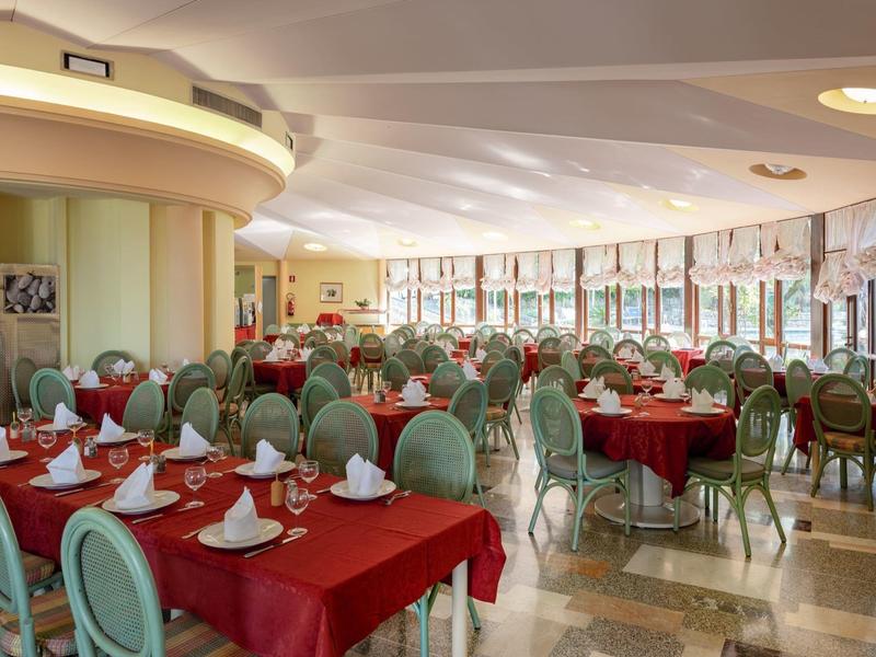 Světlá restaurace s červenými ubrusy, zelenými židlemi, velkými okny a prostřenými stoly.