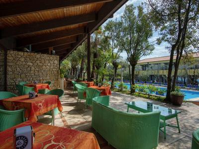 Außenbereich eines Cafés mit grünen Stühlen, orangen Tischdecken und Blick auf einen Pool und Bäume.