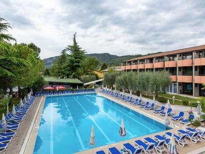 Grande piscine extérieure avec des chaises longues bleues à côté d'un bâtiment d'hôtel sous un ciel nuageux.