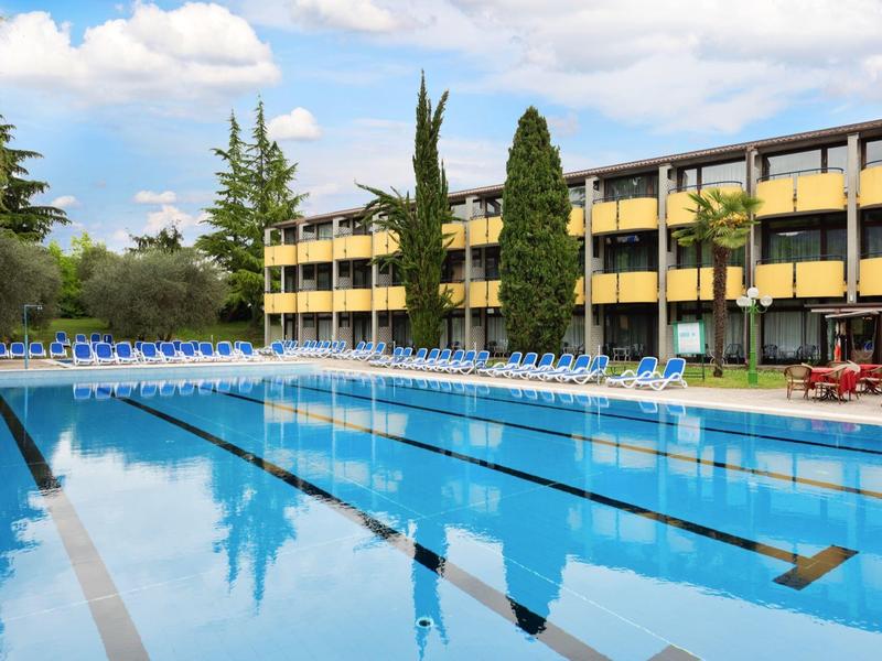 Piscine extérieure avec couloirs de nage devant un bâtiment hôtelier à plusieurs étages et parasols.