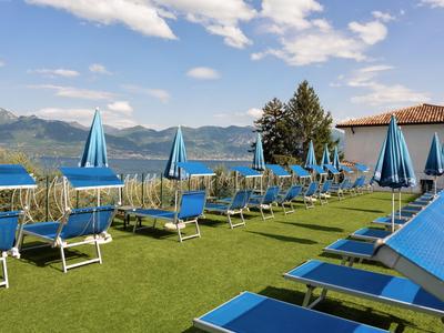Terrazza solarium con lettini e ombrelloni blu, lago di montagna sullo sfondo.