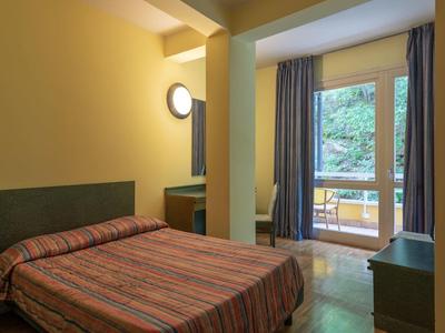 Camera d'hotel semplice con letto matrimoniale, scrivania, specchio e balcone con vista sugli alberi.