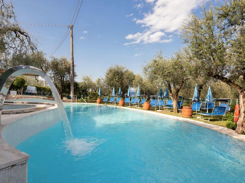 Piscina con cascata e ombrelloni blu tra gli ulivi.