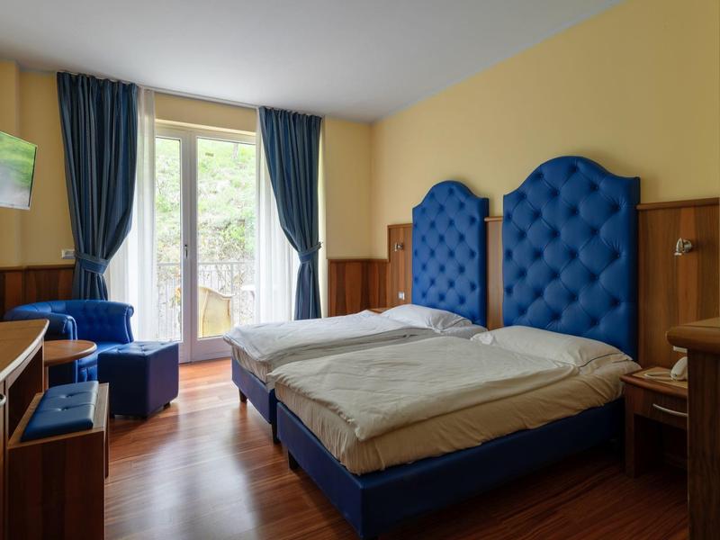 Camera d'hotel luminosa con due letti con testiere blu, una poltrona e un balcone.
