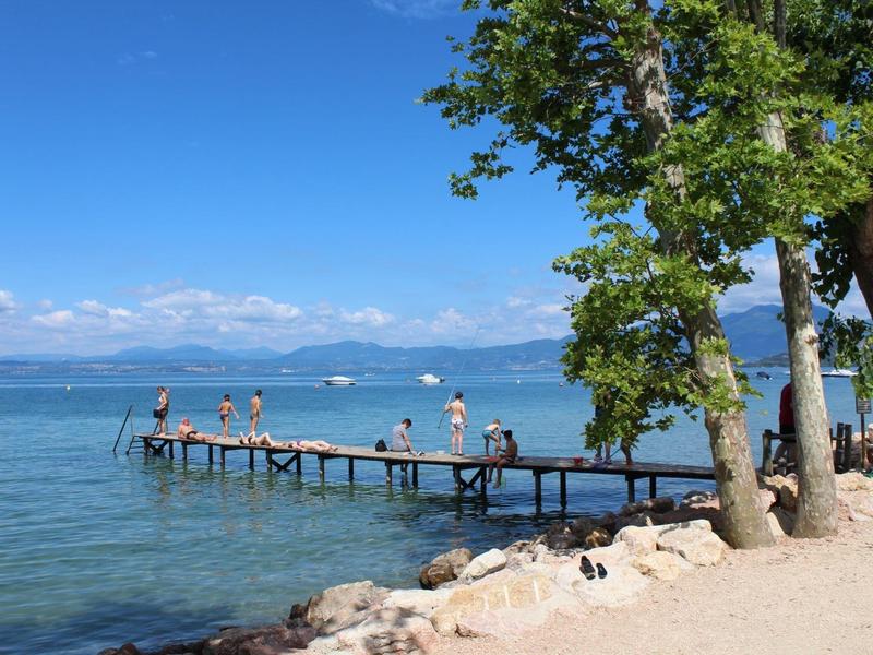 Menschen entspannen auf einem Steg am See mit klarem Wasser und blauem Himmel, Bäume am Ufer.