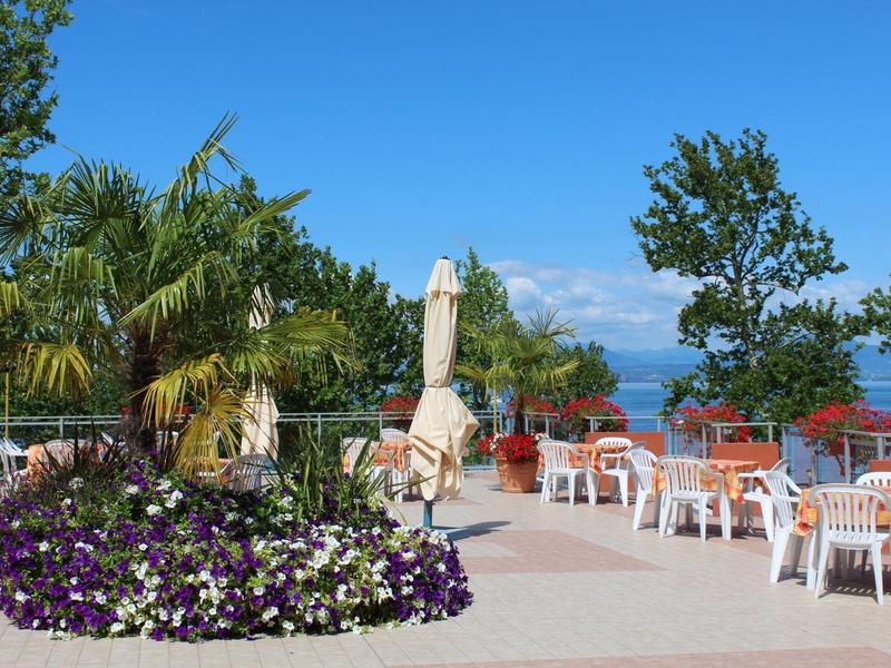 Terrasse mit weißen Tischen und Stühlen, Blumenbeet und Palmen vor blauem Himmel und Wasser.