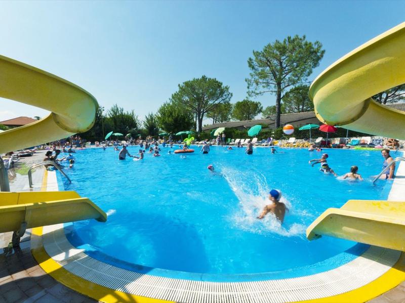 Öffentliches Schwimmbecken mit zwei gelben Wasserrutschen, vielen schwimmenden Personen und sonnigem Wetter.