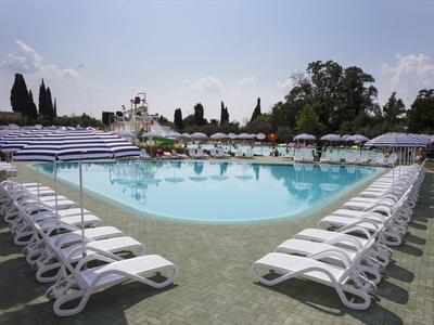 Großer, rundlicher Pool mit weißen Liegestühlen und blau-weißen Sonnenschirmen, umgeben von Bäumen.