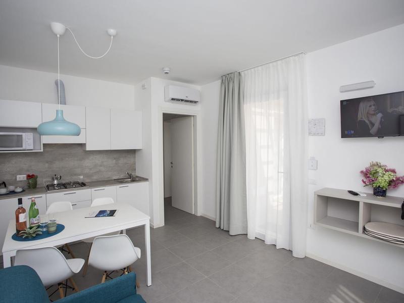 Moderner, heller Wohnbereich mit Esstisch, Küchenzeile, TV, Vorhängen und gemütlichem Sessel.