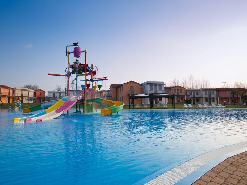 Parco acquatico per bambini con scivoli colorati accanto a una grande piscina e case vacanze.
