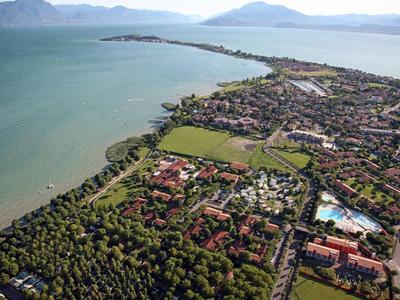 Luftaufnahme eines Sees mit angrenzendem Dorf, grünen Feldern und Bergen im Hintergrund.