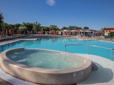 Rundlicher Whirlpool neben großem Außenpool mit Sonnenschirmen und Bäumen im Hintergrund.