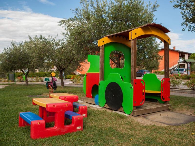 Barevné dětské hřiště s dřevěným domečkem a piknikovým stolem na travnaté ploše za slunečného počasí