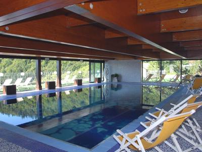 Innenliegendes Schwimmbad mit Glasfenstern, Holzbalkendecke und weißen Liegestühlen mit gelben Polstern.