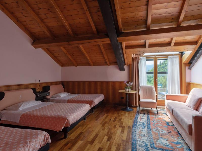 Bright room with wooden beam ceiling, two single beds, sofa, and balcony overlooking greenery.