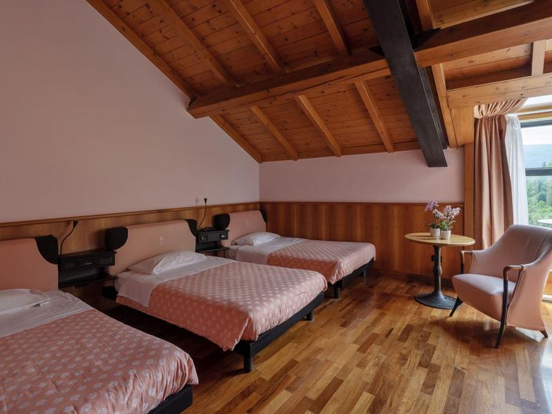 Hotel room with three single beds, wooden floor, sloped ceiling, and armchair by a window with curtains.