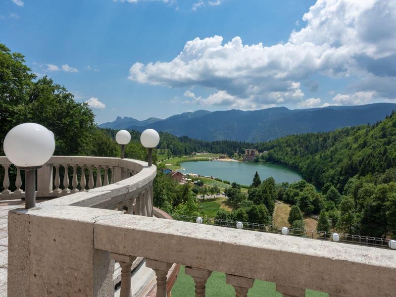 View from a terrace with globe lights overlooking a lake and forested mountains under a blue sky.