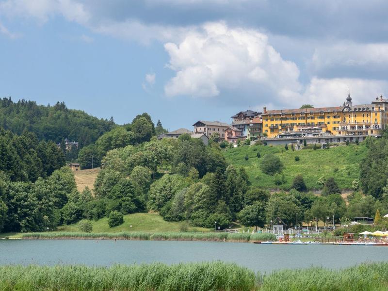 Yellow hotel situated on a wooded hill with a lake in the foreground.