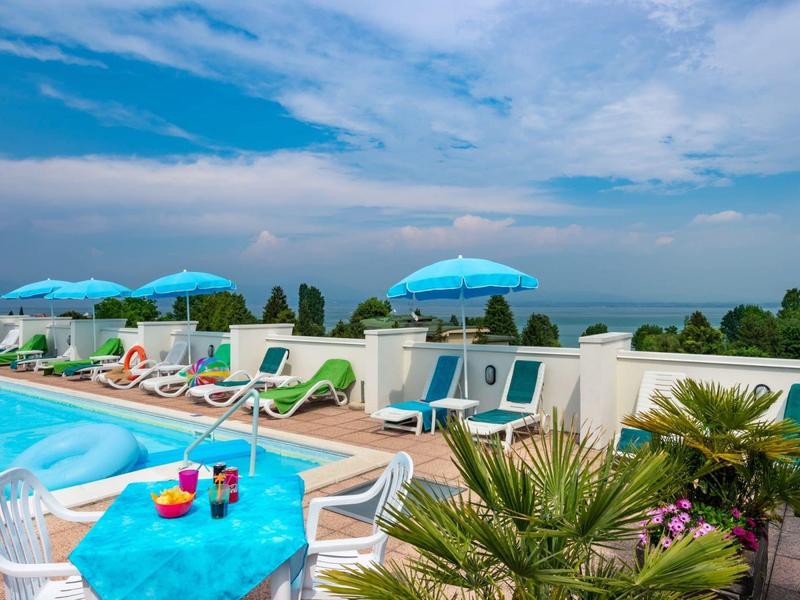 Zonnig dakterras met zwembad, ligstoelen, blauwe parasols en uitzicht op zee.