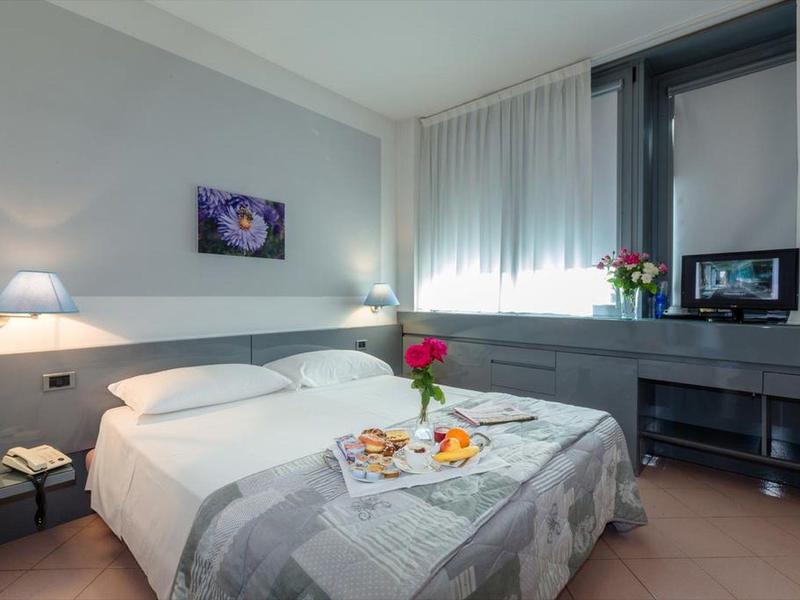 Lichte hotelkamer met tweepersoonsbed, grijze deken en ontbijtplankje met een roos.