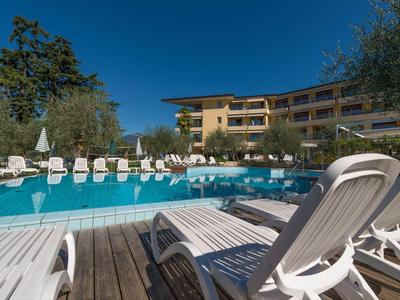 Großer blauer Pool mit weißen Liegestühlen auf Holzdeck vor gelbem Hotelgebäude unter klarem Himmel.