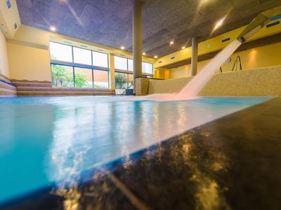 Hallenbad mit Wasserfall, großen Fenstern und hellgelben Wänden unter Decke mit blauer Mosaikoptik.