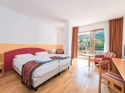 Helles Hotelzimmer mit zwei Einzelbetten, Holzboden, Balkon und Blick auf grüne Berge.