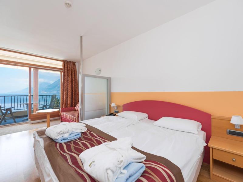 Helles Hotelzimmer mit zwei Betten, Balkon, Stuhl, Nachttisch und Deckenleuchte, helle Wände.