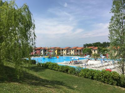 Großer Außenpool mit Liegestühlen, umgeben von grünen Bäumen und bunten Ferienhäusern.