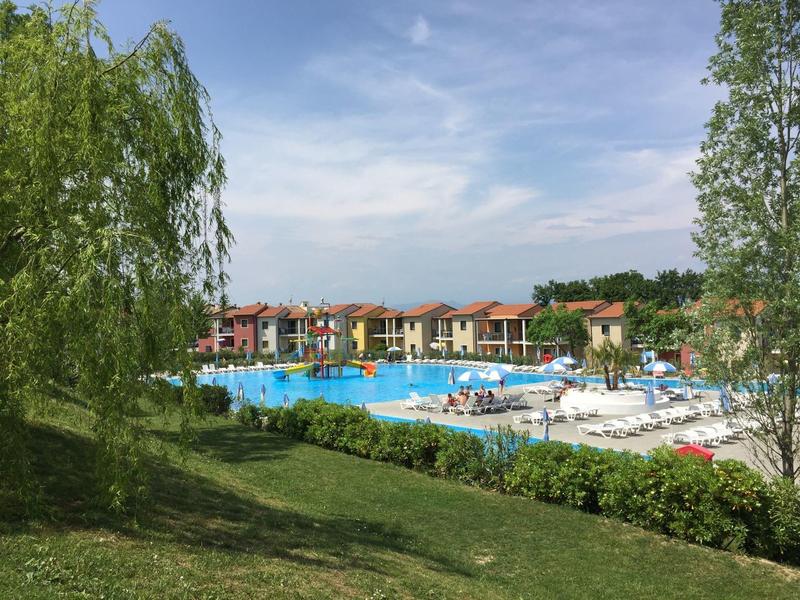 Großer Außenpool mit Liegestühlen, umgeben von grünen Bäumen und bunten Ferienhäusern.