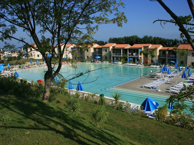 Großer Außenpool mit Liegestühlen, Sonnenschirmen und umliegenden Häusern bei klarem Himmel.