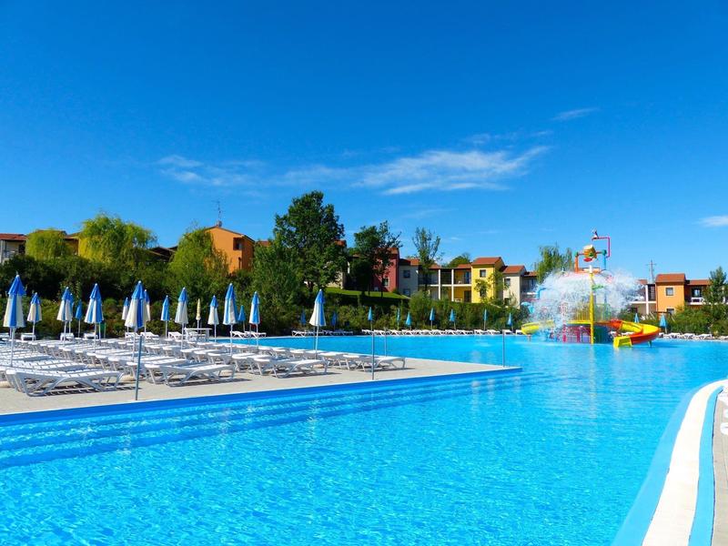 Großer Pool mit klar blauem Wasser, Liegestühlen, blauen Sonnenschirmen und bunten Häusern im Hintergrund.