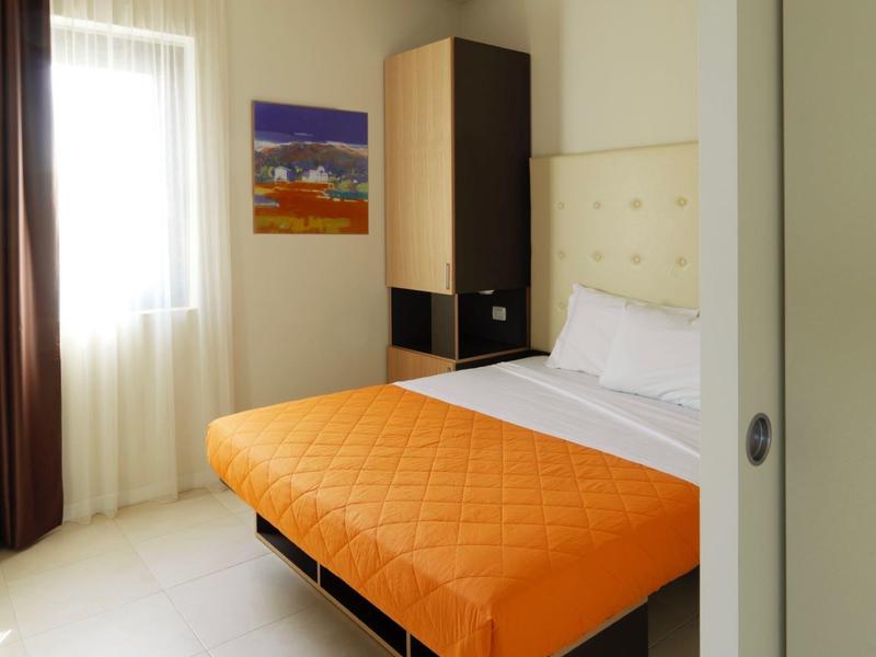 Kleines Schlafzimmer mit bett, orangefarbener Bettdecke, Schrank und Fenster mit hellen Vorhängen.