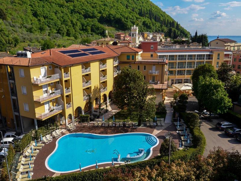 Hotel z zakrzywionym basenem zewnętrznym, leżakami i górskim krajobrazem w tle.