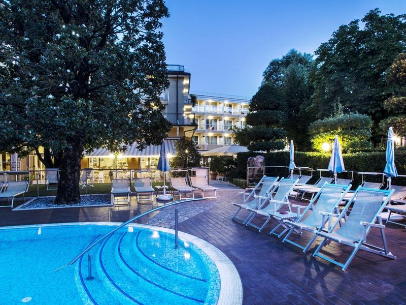 Oświetlony hotel z okrągłym basenem i leżakami o zmierzchu.