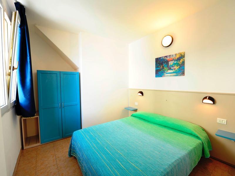 Bright hotel room with double bed, green-blue bedspread, and blue wardrobe by the window.