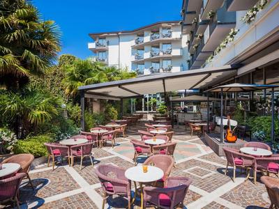 Freiluftrestaurant mit rosa Stühlen und Tischen vor einem Hotel an einem sonnigen Tag.
