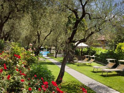 Grüner Garten mit roten Blumen, Olivenbäumen, Liegestühlen und Swimmingpool im Hintergrund.