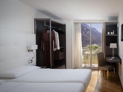 Helles Hotelzimmer mit zwei Betten, offenem Kleiderschrank und Balkon mit Bergblick.