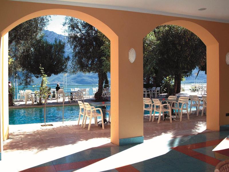 Vista a través de arcadas hacia terraza con piscina, mesas y sillas, lago y montañas al fondo.