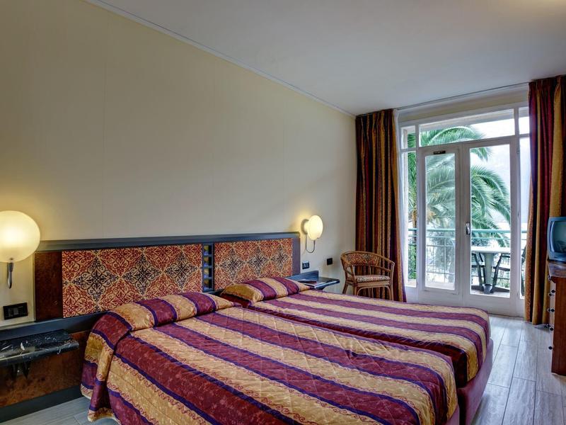 Habitación de hotel con dos camas, lámparas de mesa, balcón con vista a palmeras y cortinas marrones