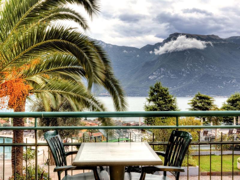 Terraza con mesa y sillas frente a palmeras y lago de montaña bajo cielo nublado.