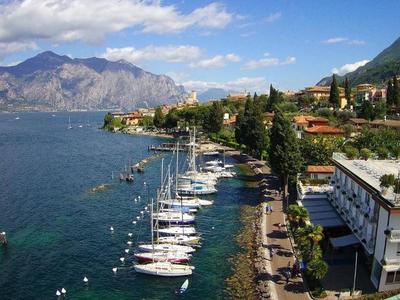 Langer Hafen mit vielen weißen Booten, blauer See, Berge im Hintergrund, Bäume und Gebäude am Ufer.