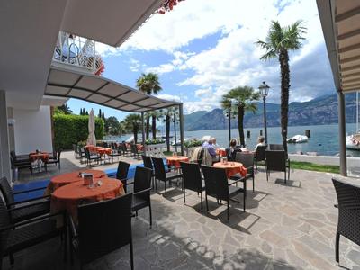 Terrasse mit Tischen und Stühlen, Blick auf Palmen und See unter blauem Himmel.